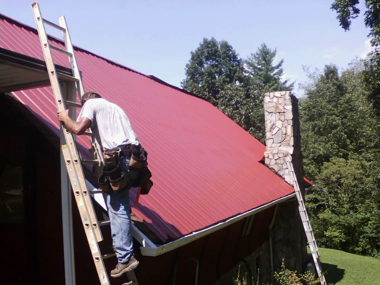 Roofer on a ladder beside a red metal roof and stone chimney — Soffit & Fascia Repair by Roofing Brothers in Lyndeborough